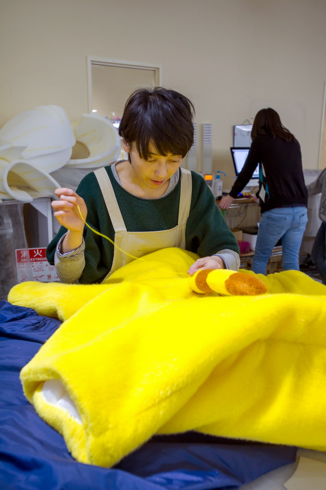 Woman sewing bright yellow fabric by hand at a workstation in a mascot suit factory.