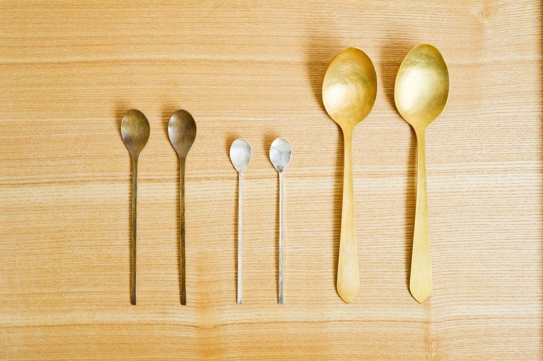Six spoons of varying sizes arranged on a light wooden surface, including smaller metallic spoons and larger gold-toned spoons.