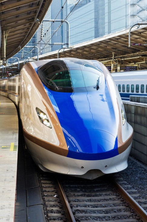 A white, blue and brown Hokuriku Shinkansen bullet train at a station platform under a metal canopy.