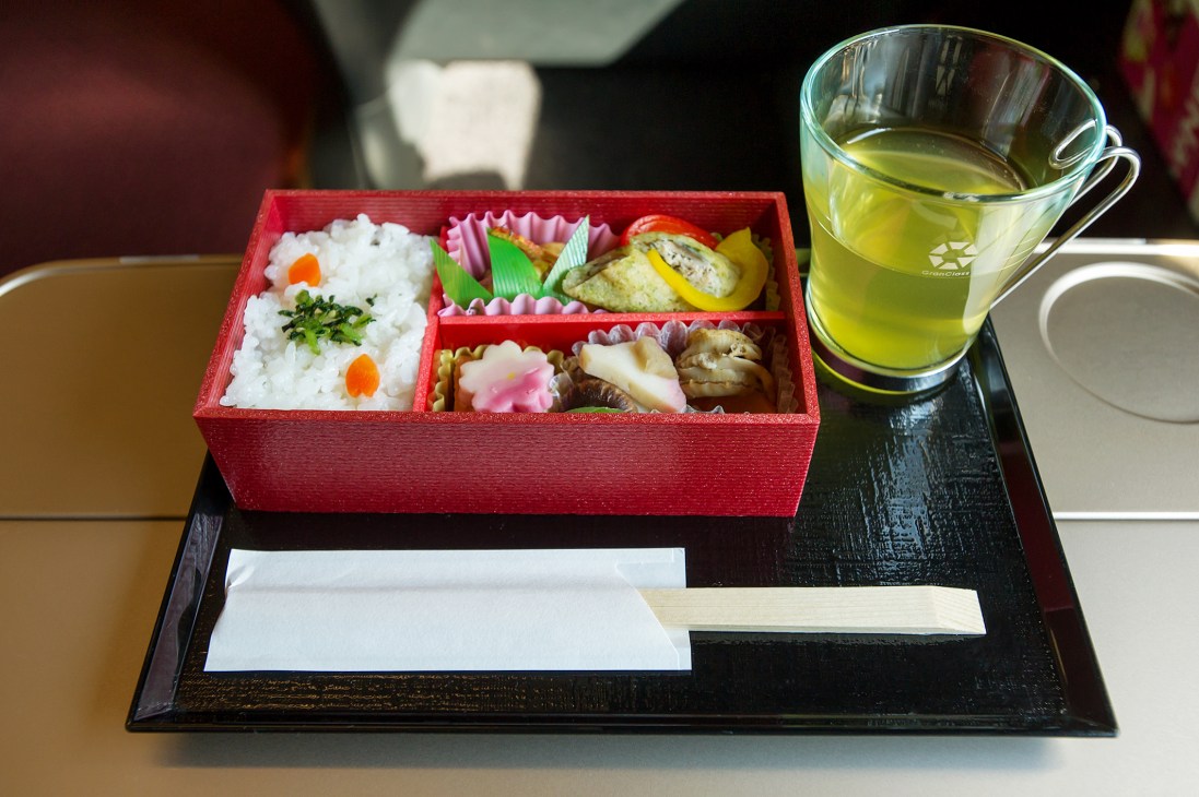 A red lacquered bento box containing rice, vegetables, and fish on a dark tray with green tea in a glass cup.