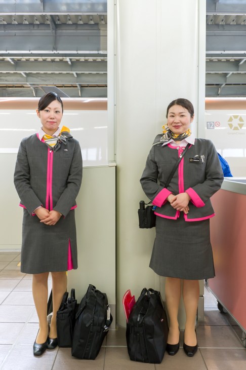 Two female train attendants in grey and pink uniforms standing in a Shinkansen train carriage.