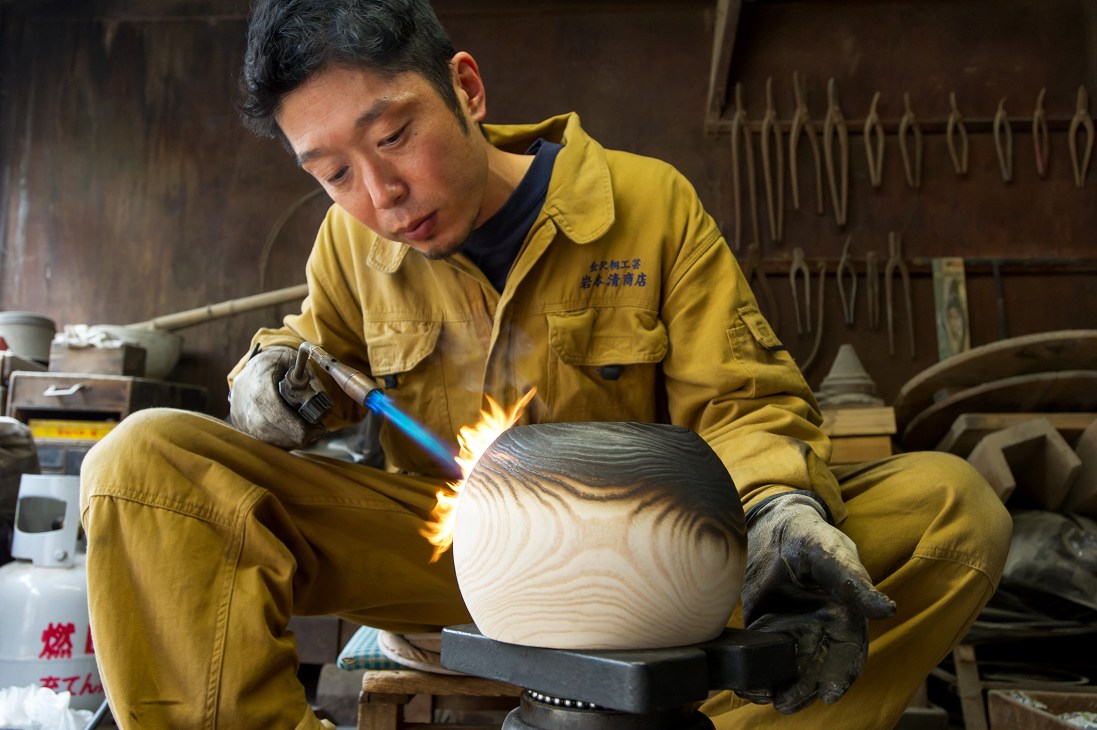 Craftsman Kensuke Uchida torches paulownia wood in yellow work clothes at Iwamoto Kiyoshi Shouten workshop.
