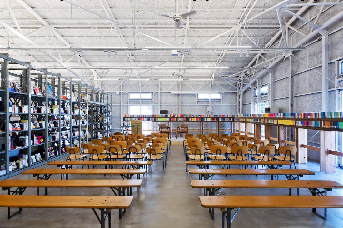 La Kagu's warehouse lecture space with wooden tables, orange chairs, industrial ceiling, and colorful book spine display.