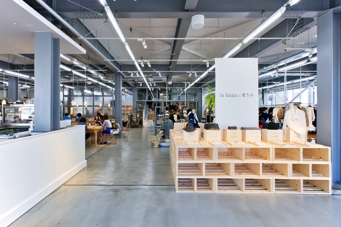 La Kagu bookstore interior with wooden shelving displaying books and a white sign with Japanese characters, amid modern warehouse space with pendant lights and customers browsing.