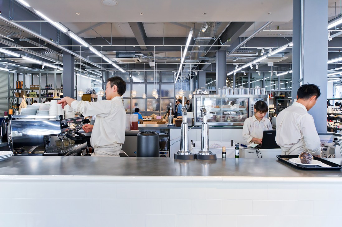 Café counter with baristas in white uniforms preparing drinks in a modern, minimalist space with industrial lighting.