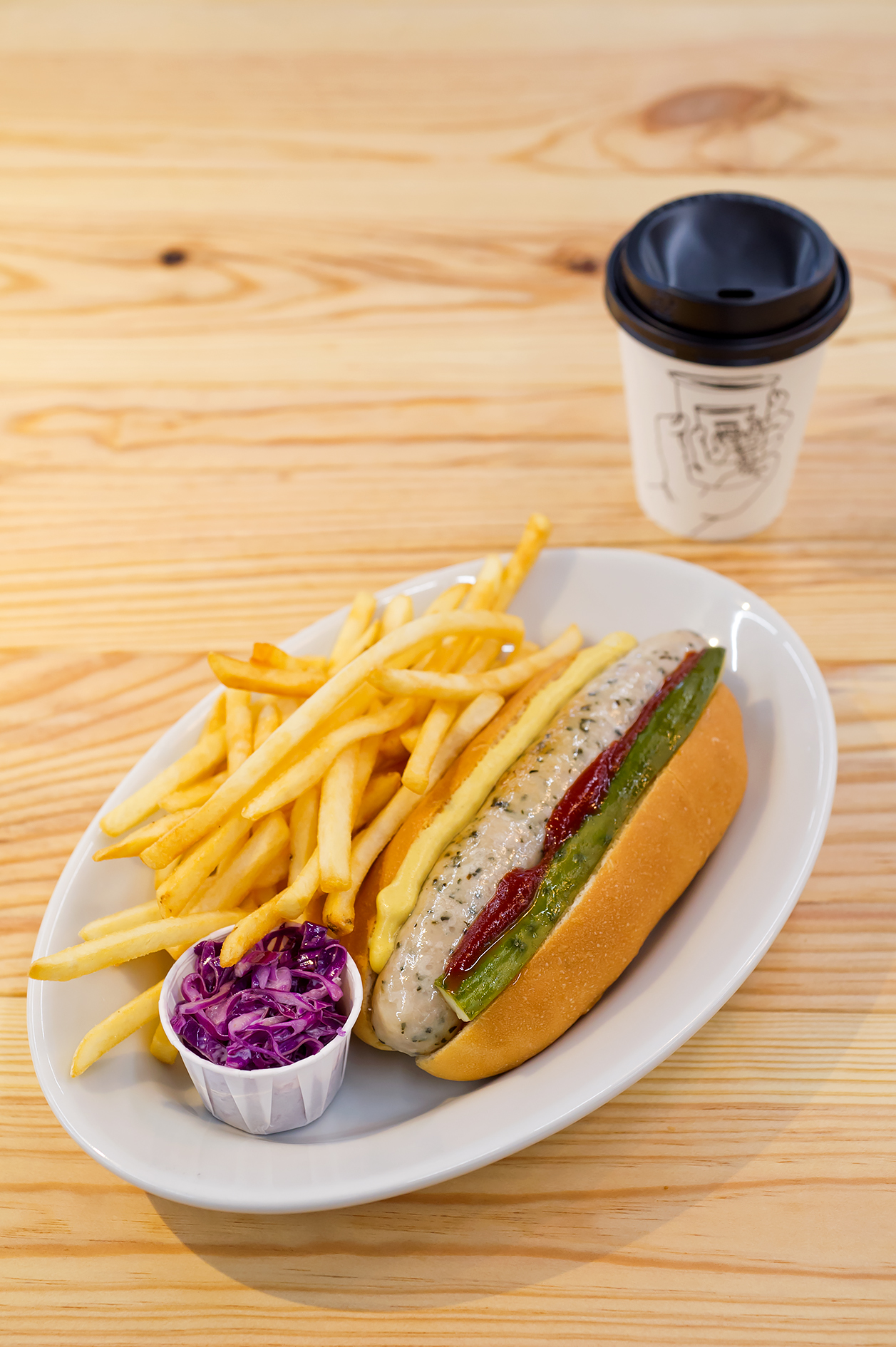 Gourmet hot dog with toppings and fries on a white plate with a coffee cup on wooden table.