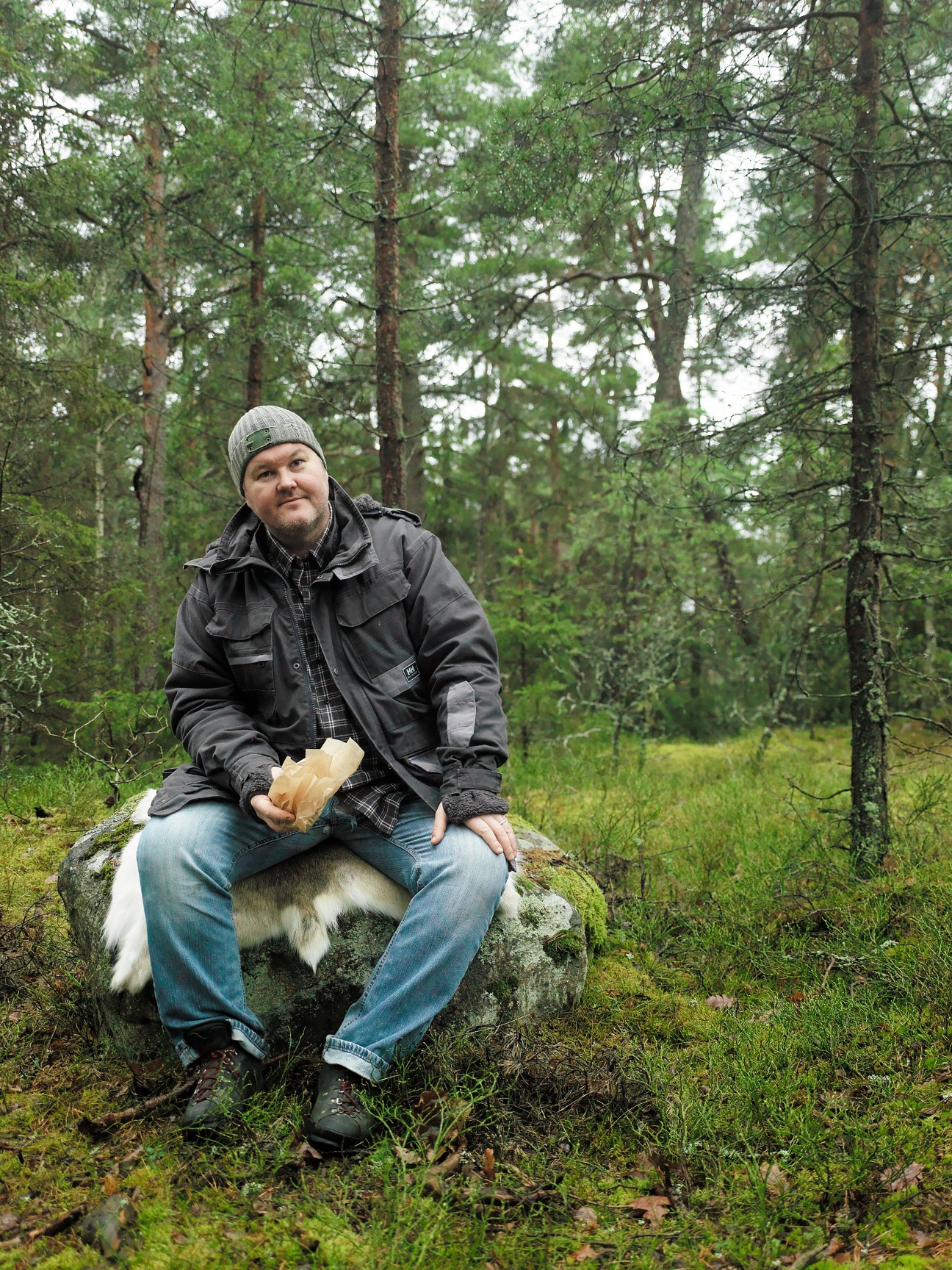 Chef Mathias Dahlgren sits on a moss-covered rock in a forest clearing, holding bread and wearing a beanie and black jacket.