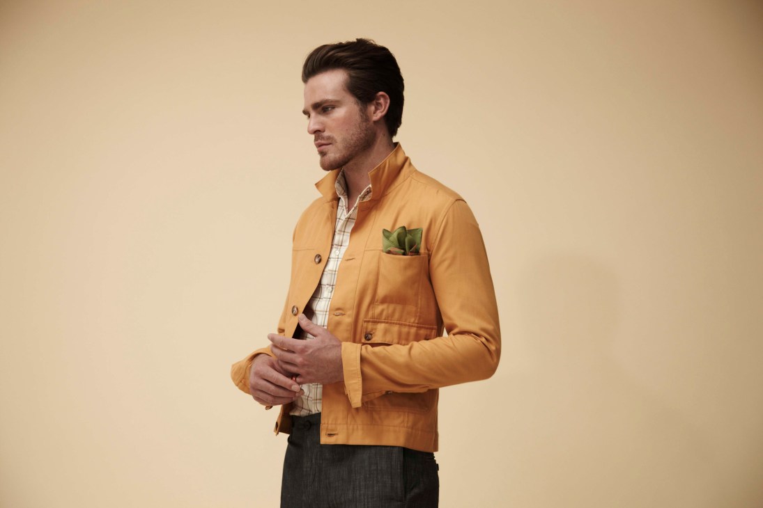Man wearing a mustard yellow jacket with checkered shirt and succulent pocket square against neutral background.