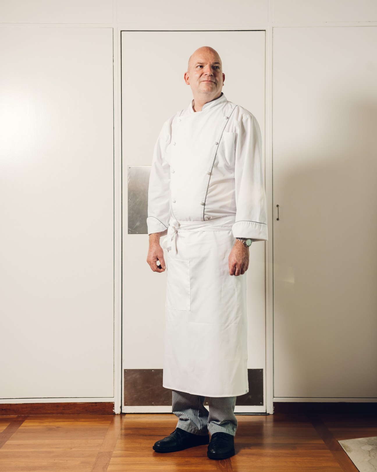 Chef Jens Top Fisker stands in white chef's coat and checkered trousers in a modern kitchen.