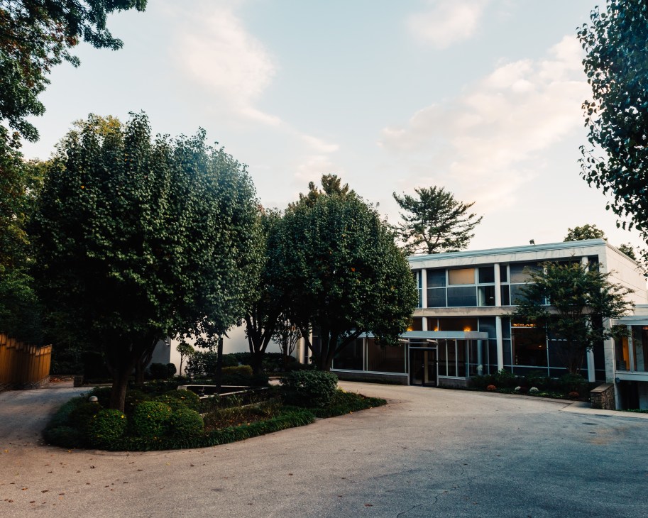 Modern white building with large windows and blue-green roof surrounded by tall trees.