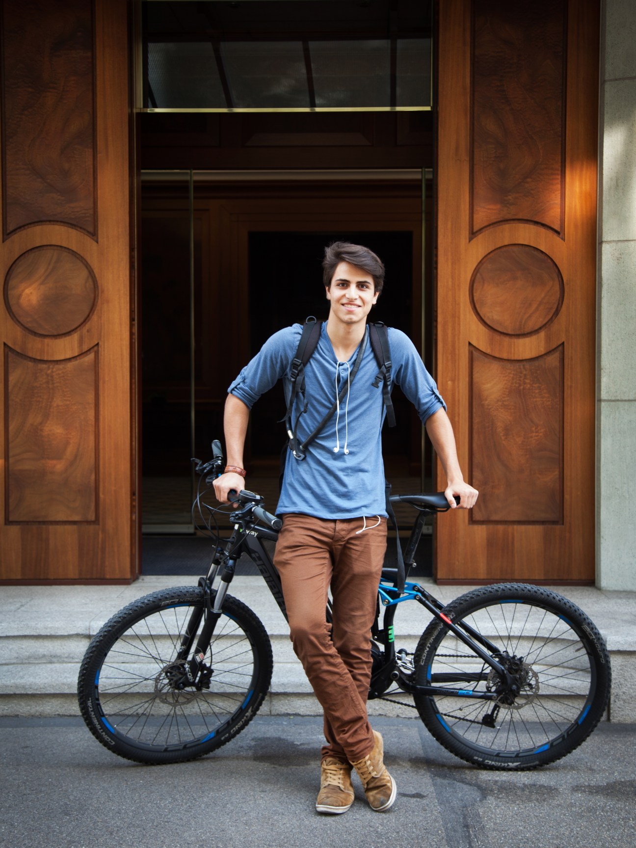 Young man holding a black and blue electric bike in front of ornate wooden doors on a Zurich street.