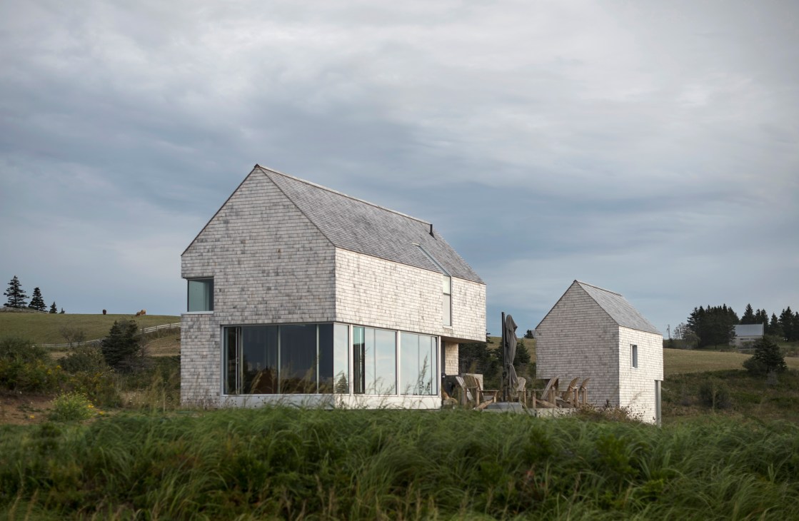 Modern cedar-shingled house with large windows set in rural landscape under cloudy sky