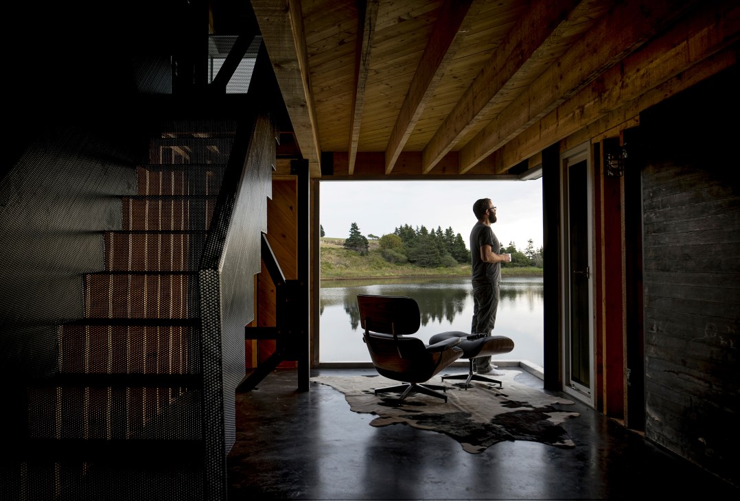 Man in dark clothing standing on deck overlooking lake with exposed wooden ceiling beams and modern chair