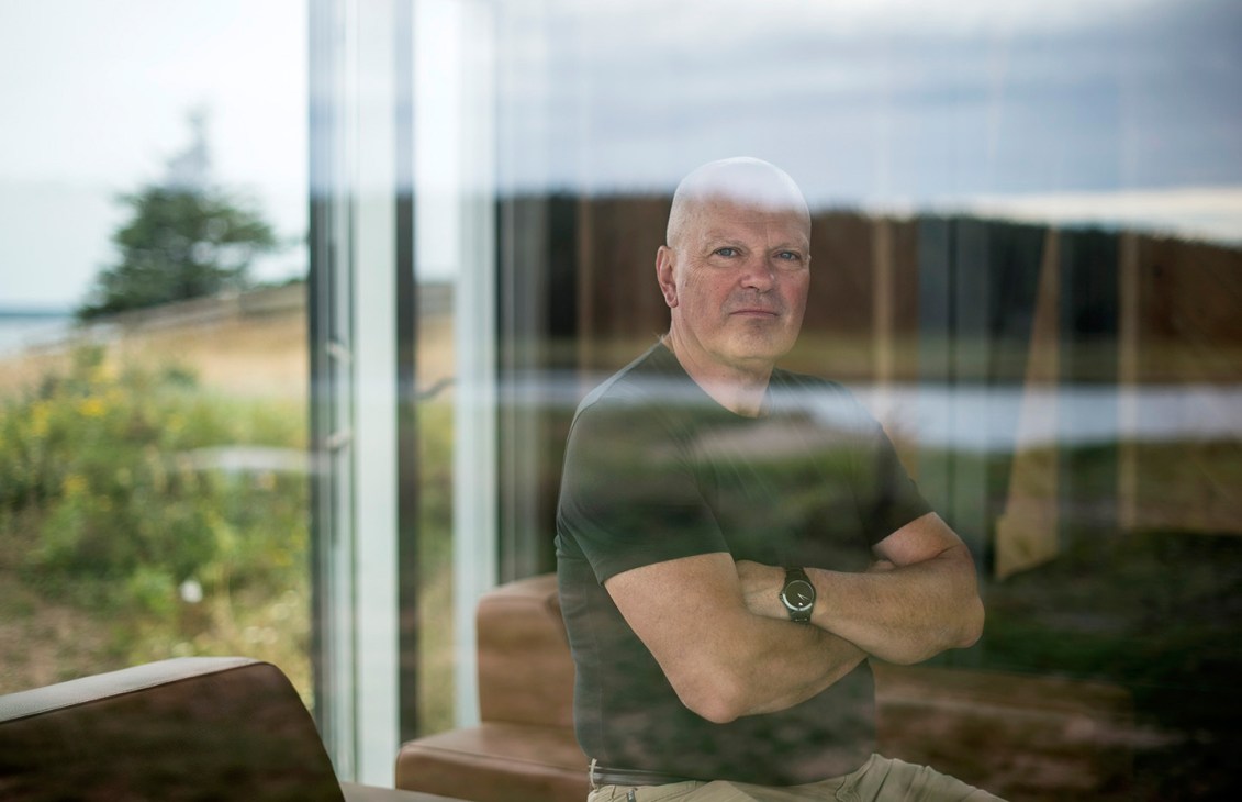 Brian MacKay-Lyons stands with crossed arms by a large window overlooking a modern building exterior