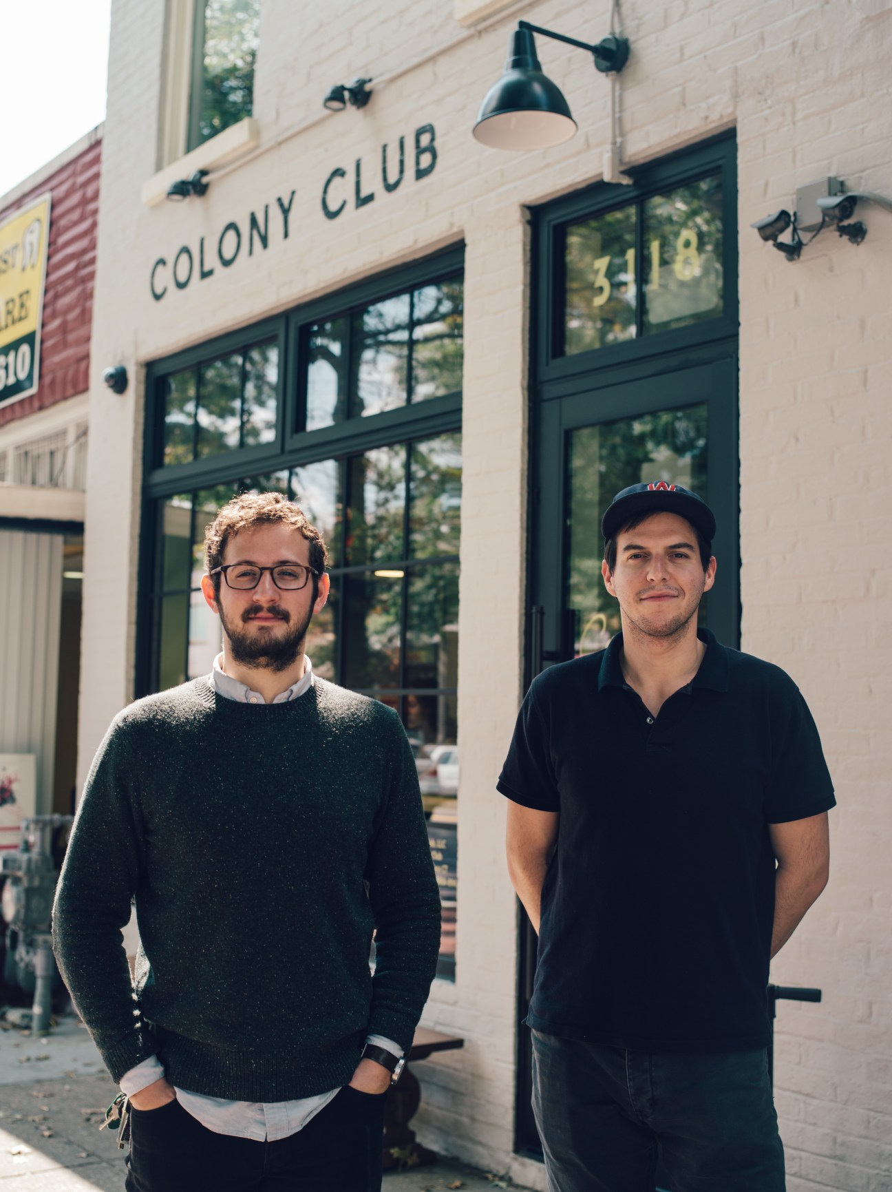 Two men stand outside Colony Club coffee shop in Washington, DC, with dark-framed windows and pendant lights visible behind them.