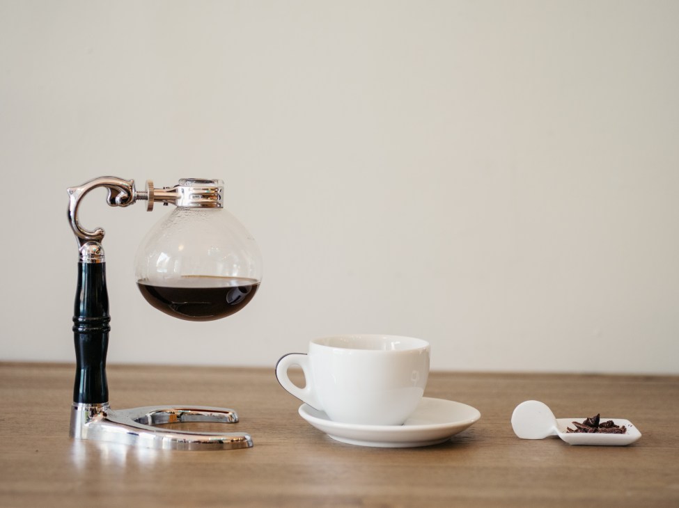 A vintage coffee siphon with dark liquid next to a white cup and saucer on a wooden surface.