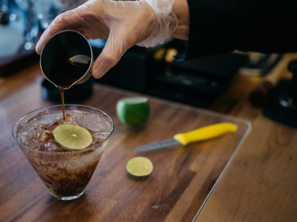 A bartender pours dark spirit into a glass with ice and lime slice on a wooden counter.