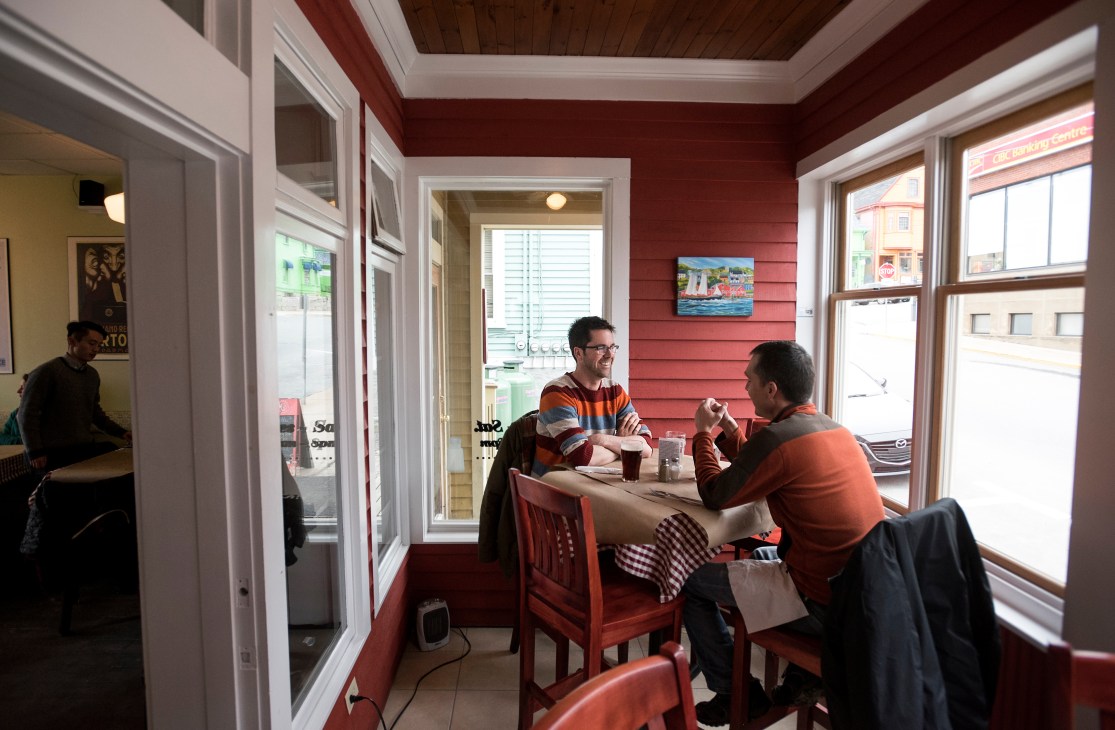 Two men dining together at a table inside Salt Shaker Deli in Lunenburg, with red walls and large windows.