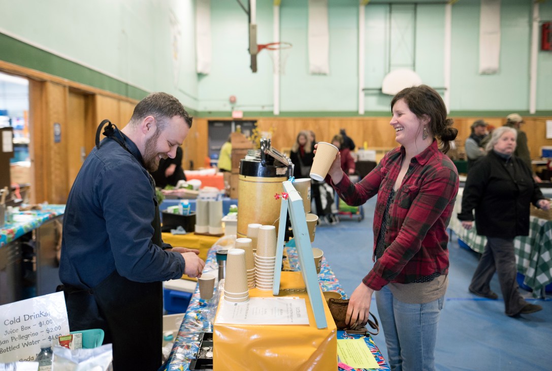 Woman serving a beverage to a man at an indoor community market event in a gymnasium.