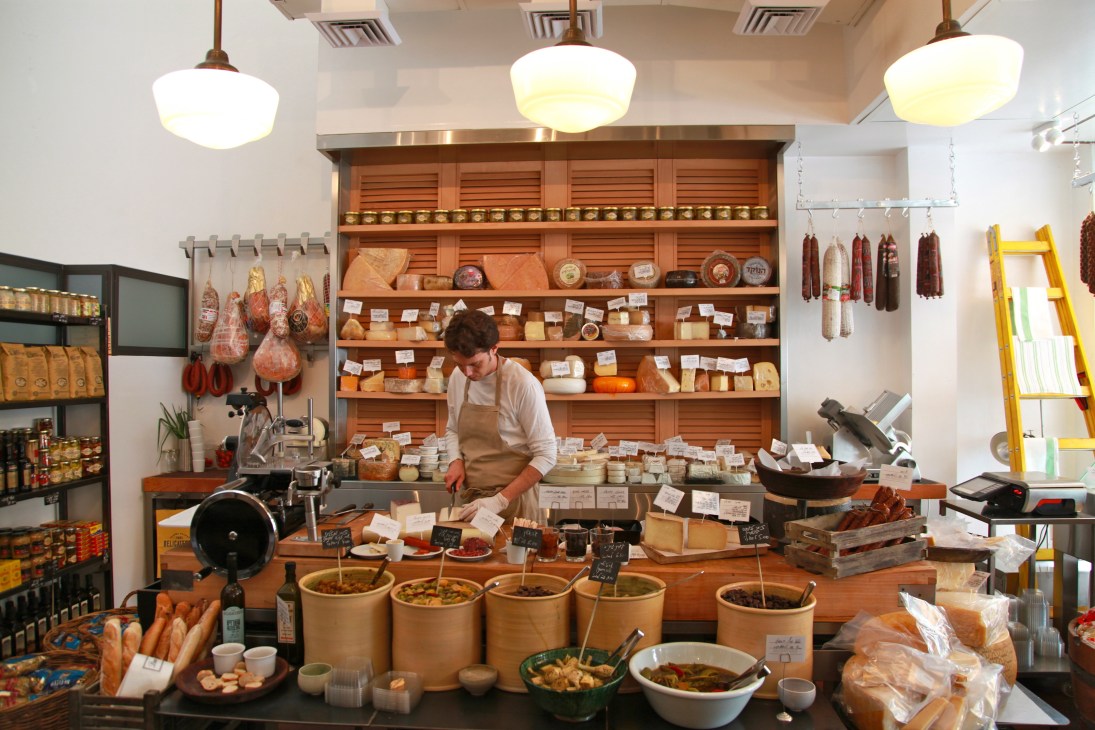 Interior of Delicatessen deli counter with staff member behind wooden shelving displaying cured meats, cheeses, and gourmet products.