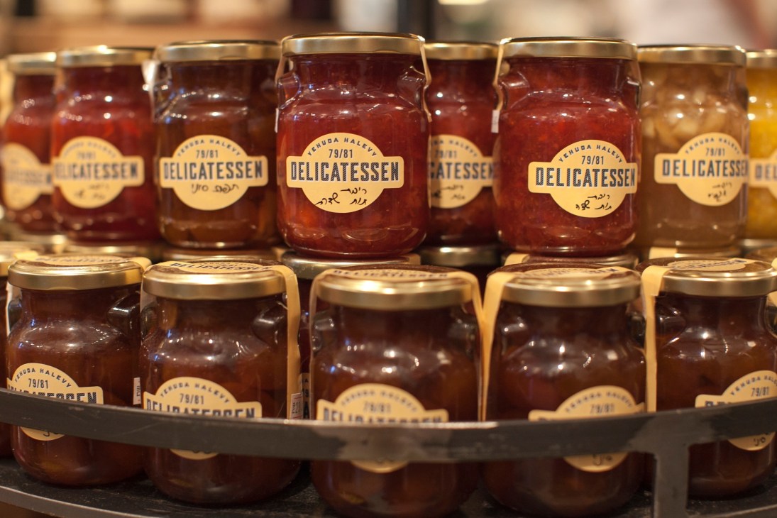 Rows of glass jars filled with preserved vegetables and jams labeled 'Delicatessen' with gold lids.