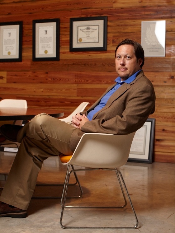 Dale Riser of Beard + Riser Architects sits in a modern chair against a wooden wall with framed certificates.