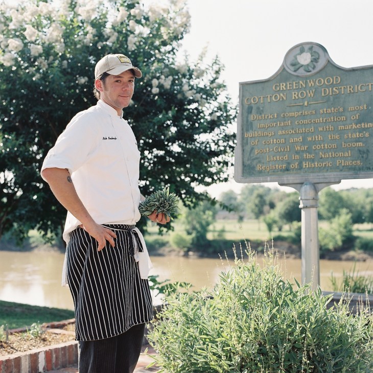 Chef Nick Seaburgh standing outdoors by the Greenwood Cotton Row District historical marker, holding fresh herbs.