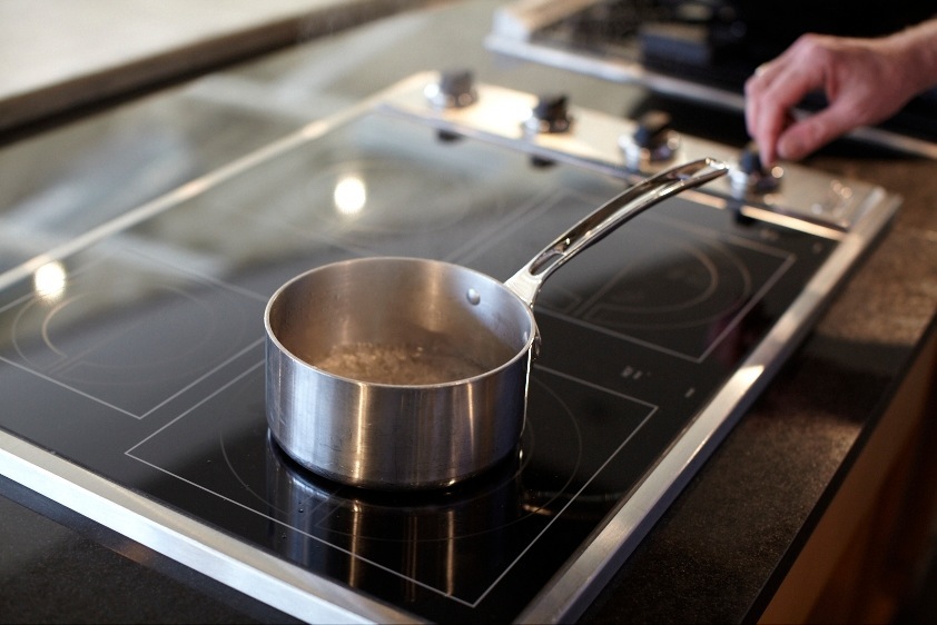 Hand hovering over a stainless steel pot on Viking's All-Induction Cooktop with black surface and control knobs.