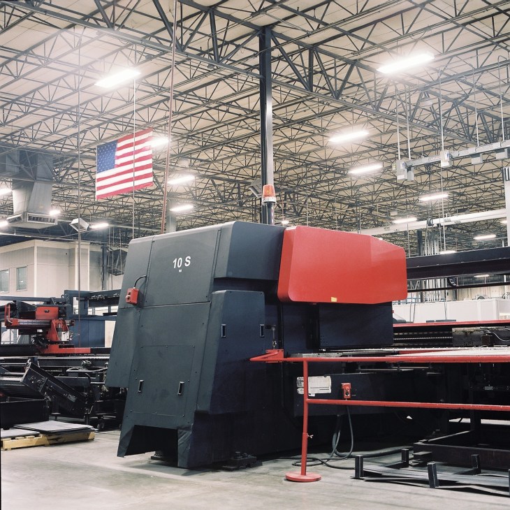 Industrial manufacturing machine with red and black components in a large factory warehouse with American flag.