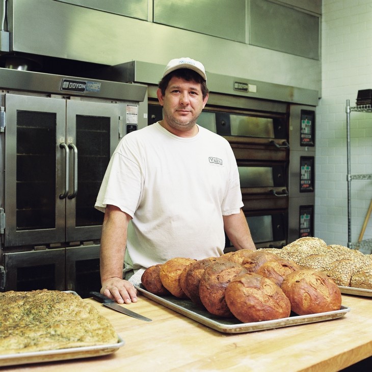 Baker in Viking-branded shirt displays fresh bread and seeded loaves at the cookery school kitchen.