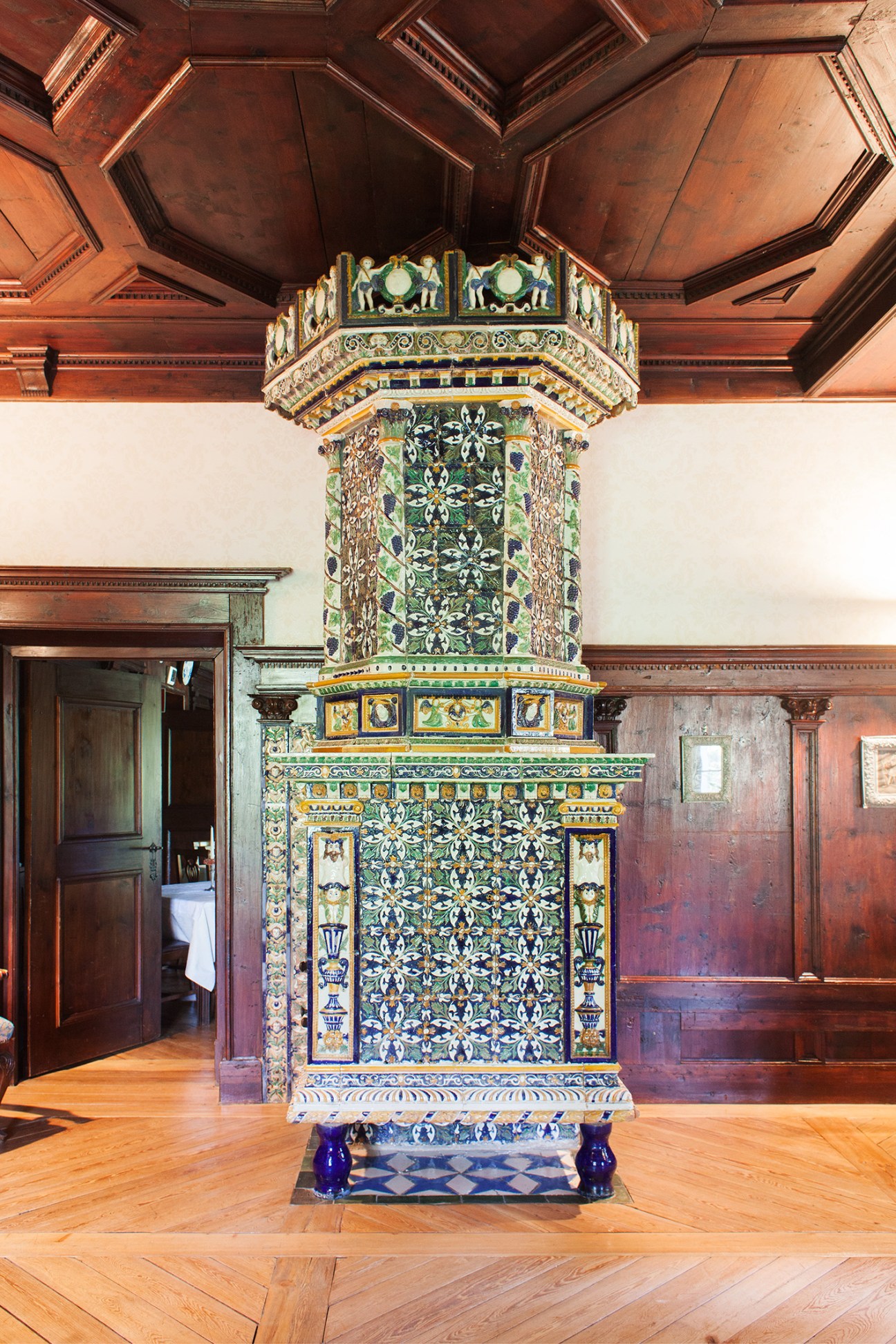 An ornate 17th-century Austrian furnace covered in colorful glazed majolica tiles featuring geometric and floral patterns, with a decorative crown-like top beneath a wooden ceiling.
