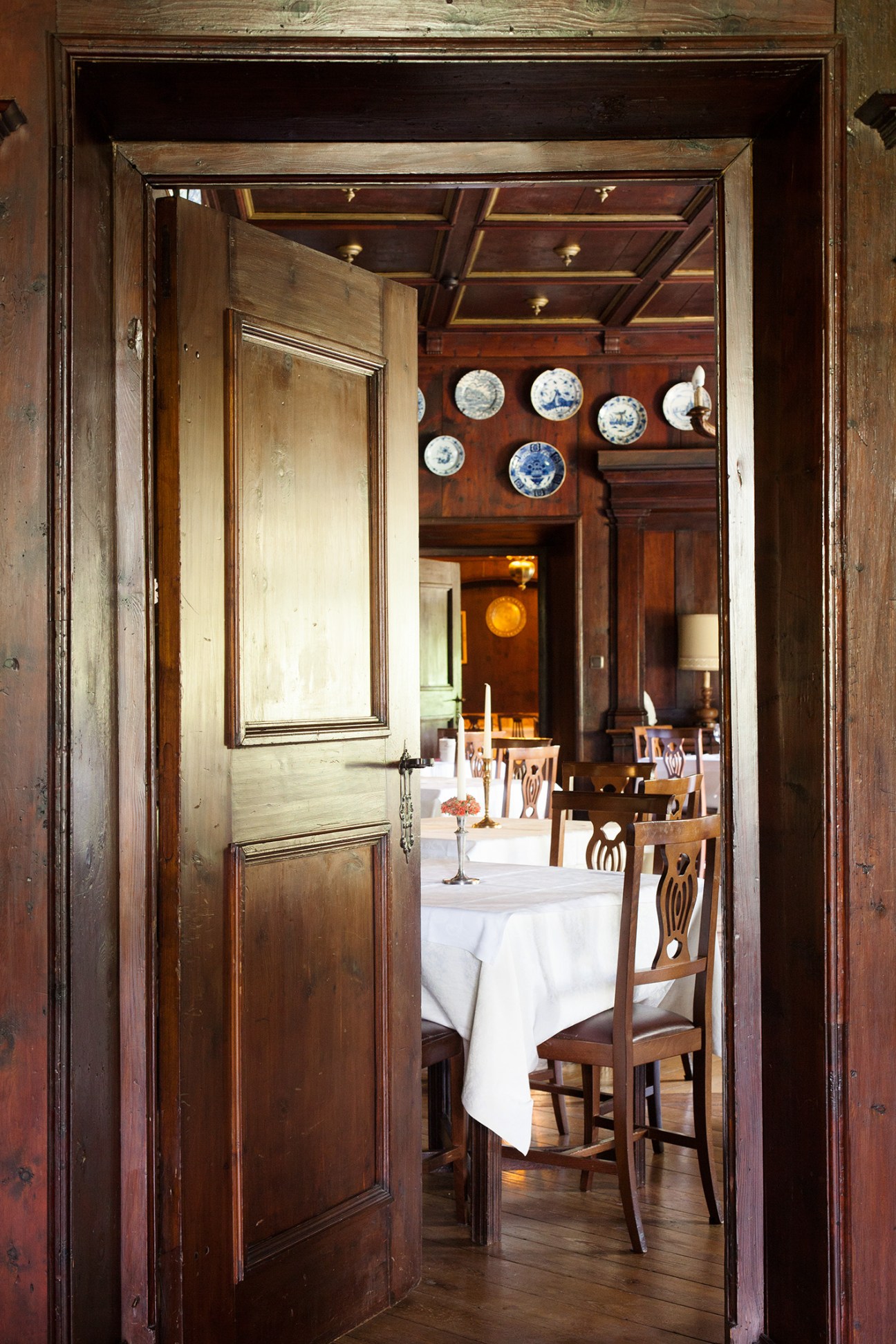 View through wooden doorway into dining room with porcelain plates displayed on wall above wooden chairs and white tablecloths.