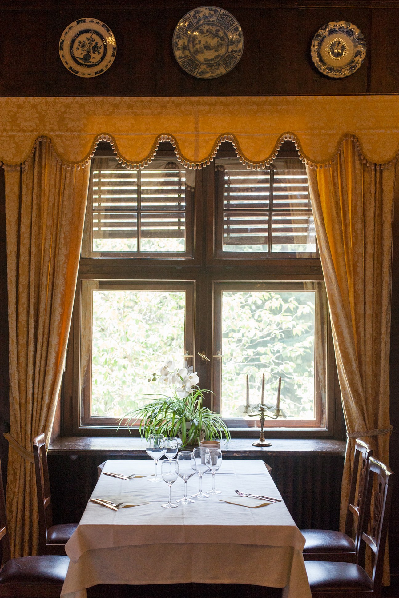Dining room with set table, ornate porcelain plates on dark wall, and large windows with golden curtains overlooking gardens.
