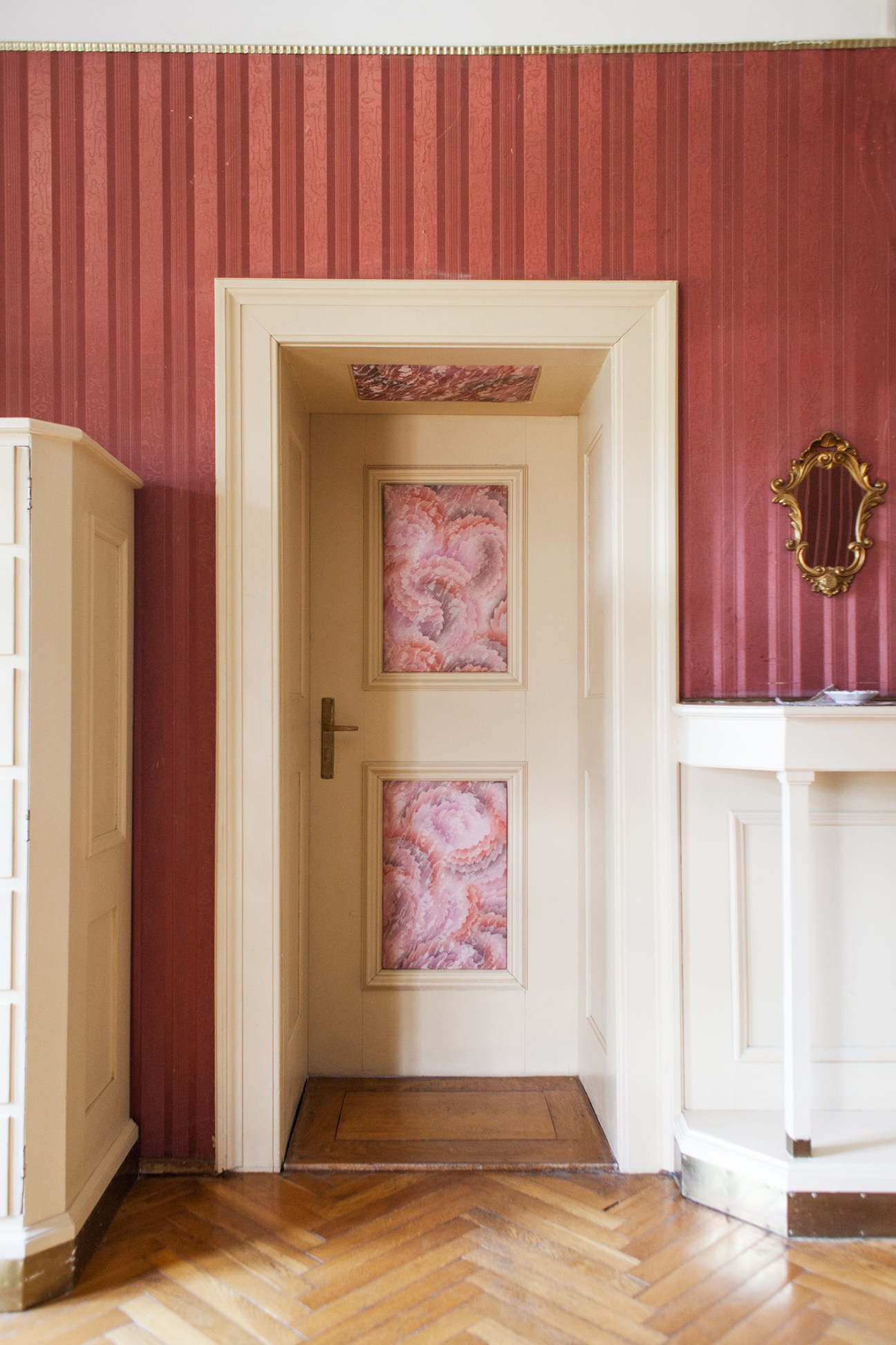 Cream-framed doorway with floral artwork panels leading into a guest room, flanked by red damask walls.