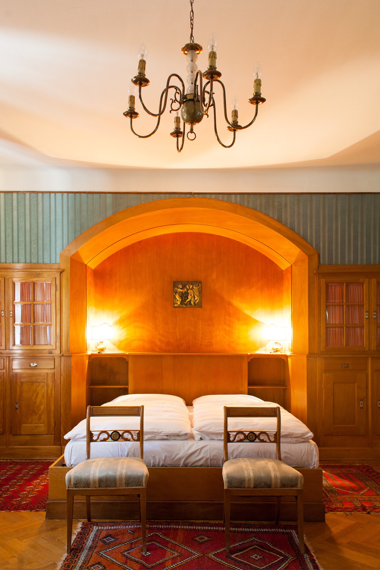 A bedroom at Castello Stifterhof with an ornate wooden bed, glowing orange alcove headboard, brass chandelier, and period furnishings.