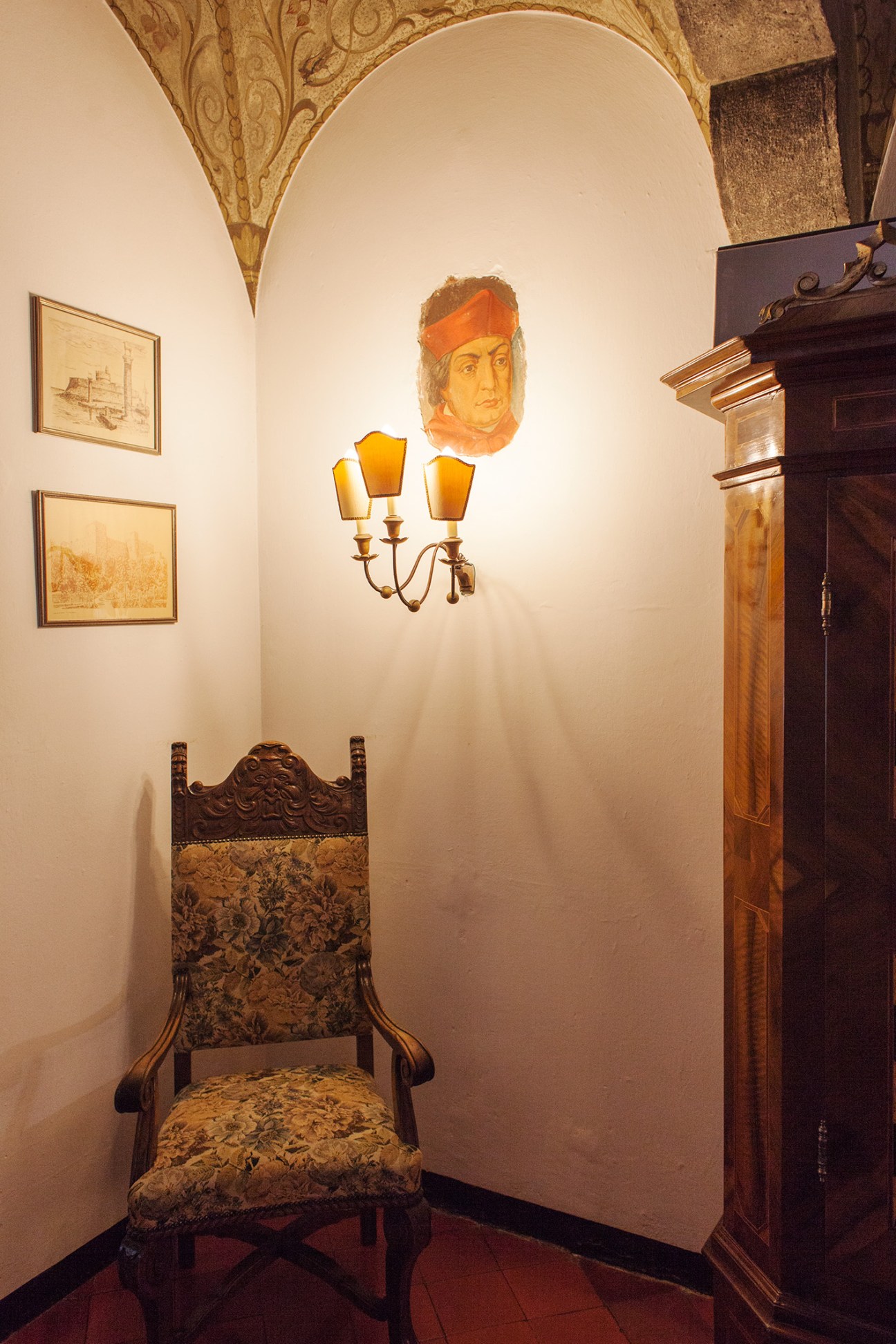 Ornate wooden chair with fabric seat beneath a gold wall sconce and portrait painting in arched alcove.