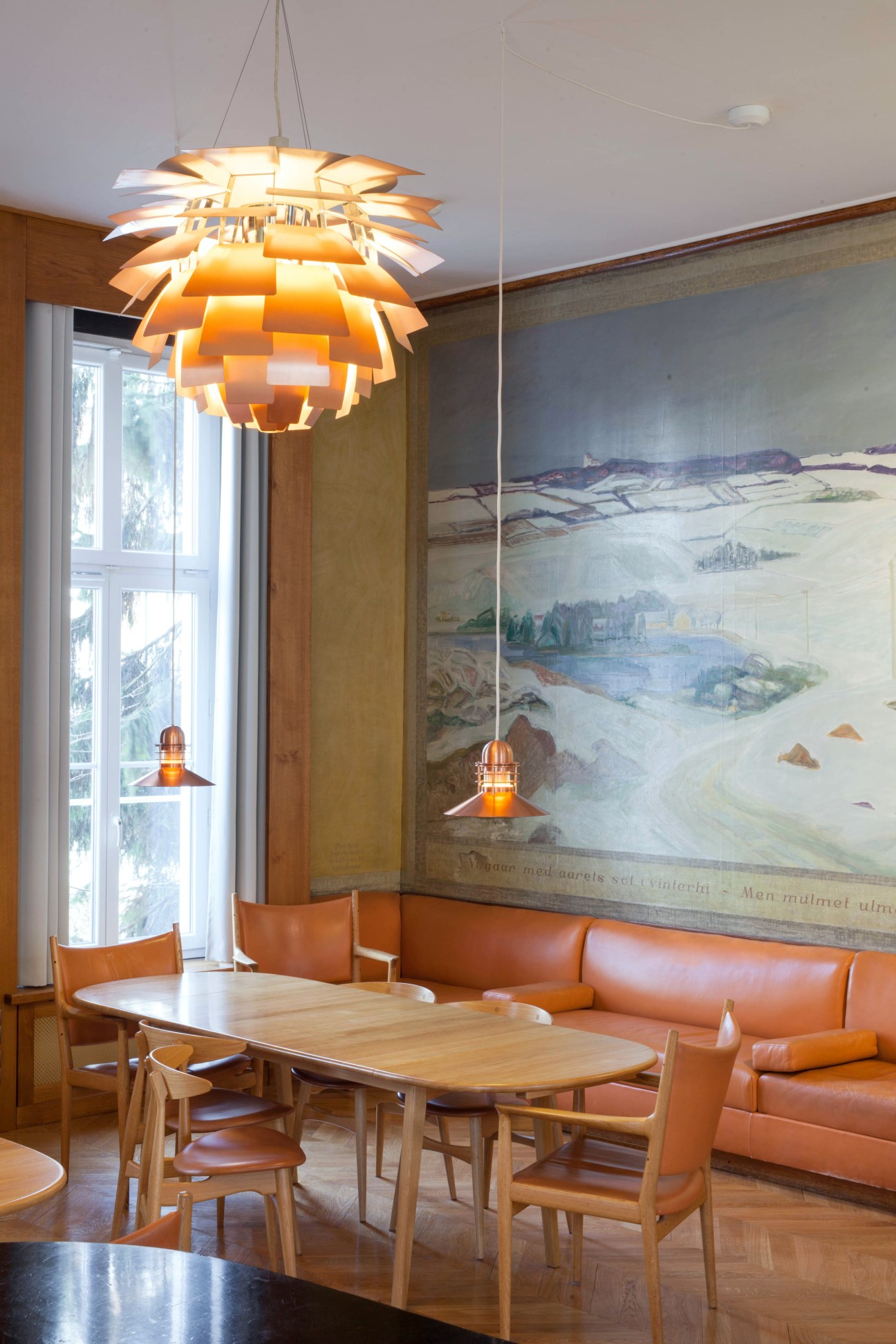 Orange pendant lamp hanging above a wooden dining table with leather chairs in a bright common room with landscape murals.