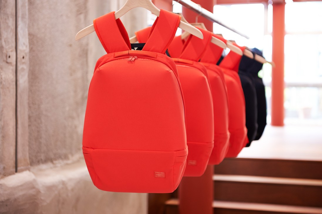 Red backpacks displayed on wooden hangers in a minimalist retail space with concrete walls and red structural elements.