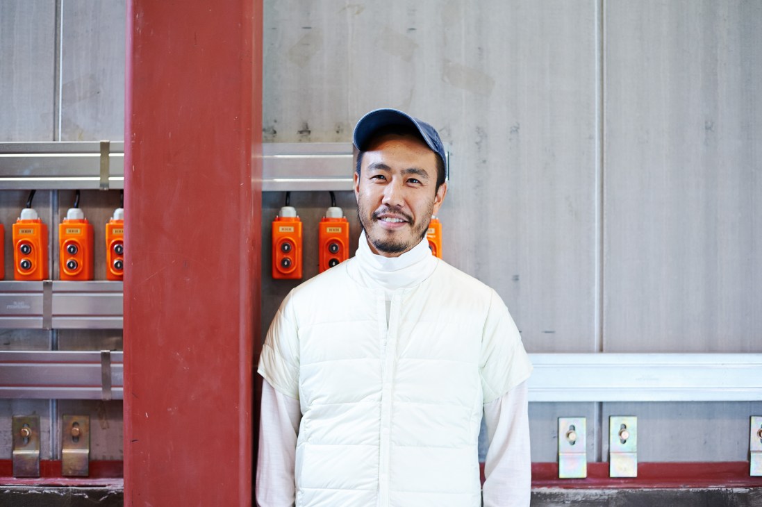 Man wearing white puffer vest and navy cap in front of Descente Blanc storefront with orange bottles displayed.
