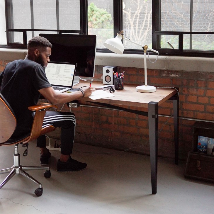 Man working at wooden desk with laptop and Floyd modular furniture legs in industrial loft space.
