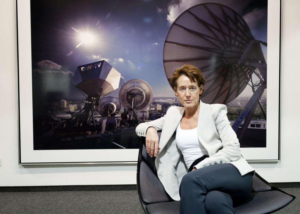 Dagmar Engel, editor-in-chief of Deutsche Welle, sits in front of a photograph showing broadcasting satellite dishes and equipment.