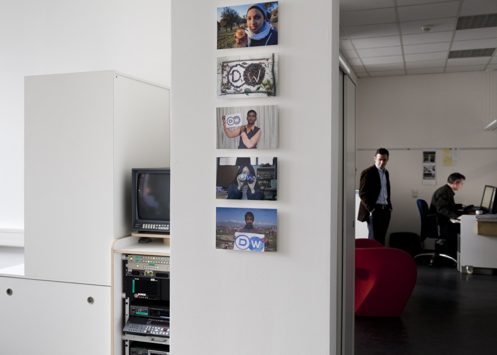 Deutsche Welle office wall displaying photographs of staff members from around the world holding DW logos.