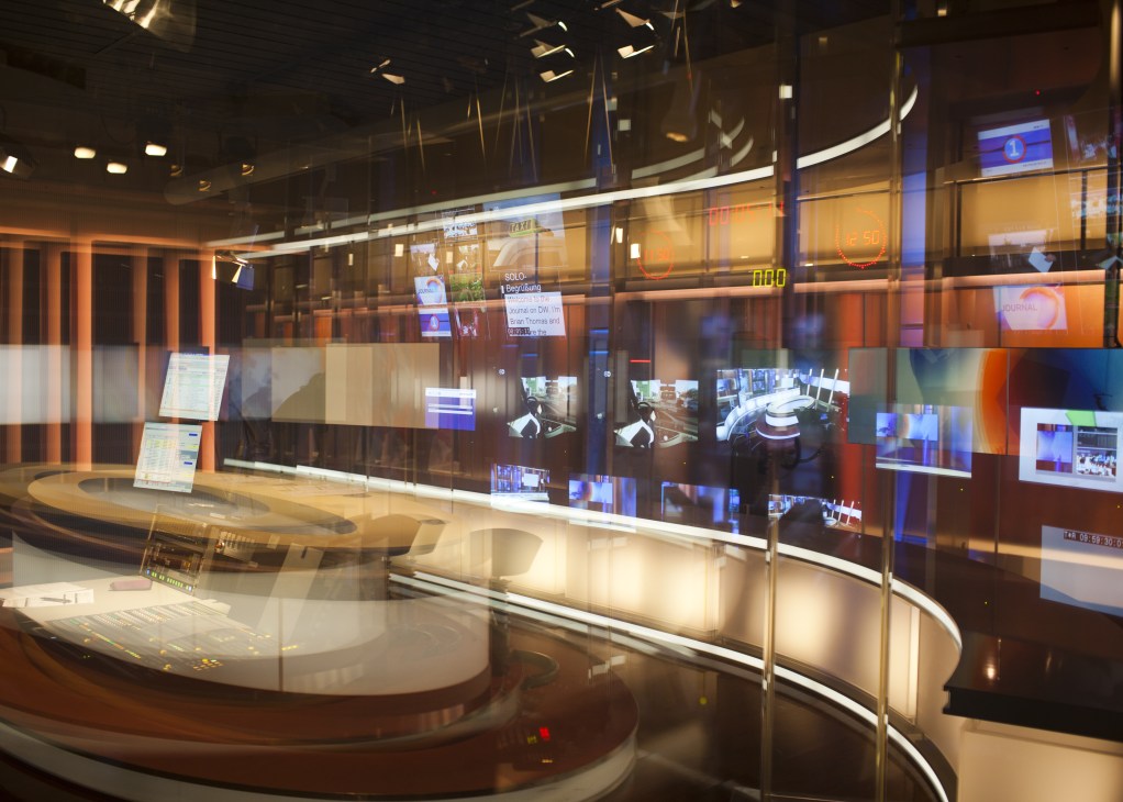 Empty Deutsche Welle television studio with curved desk, multiple monitors, and orange and blue lighting before broadcast.