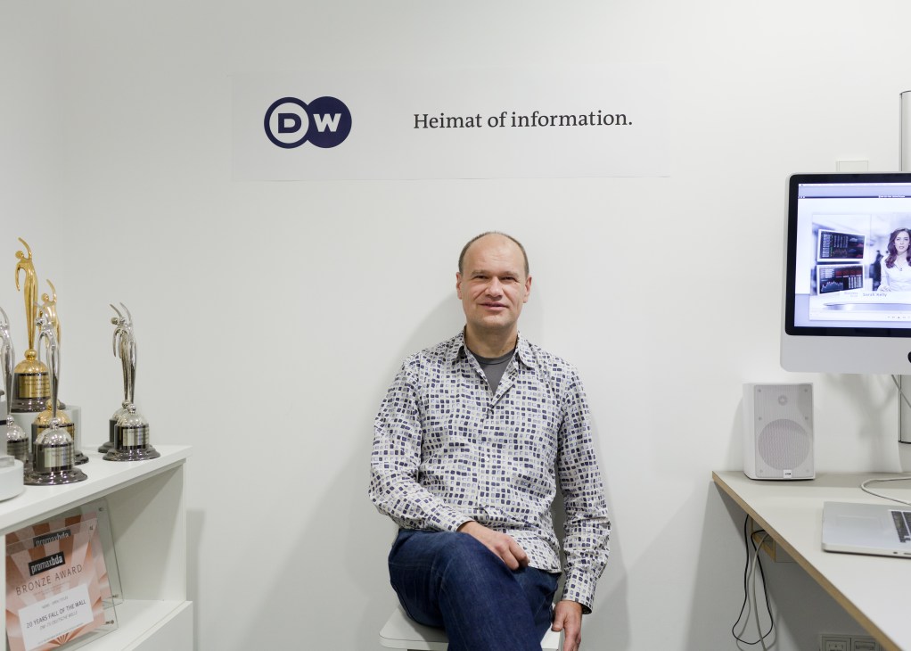 Holger Zeh, creative director of graphic design at Deutsche Welle, sits in an office with the DW logo and 'Heimat of information' tagline on the wall behind him.