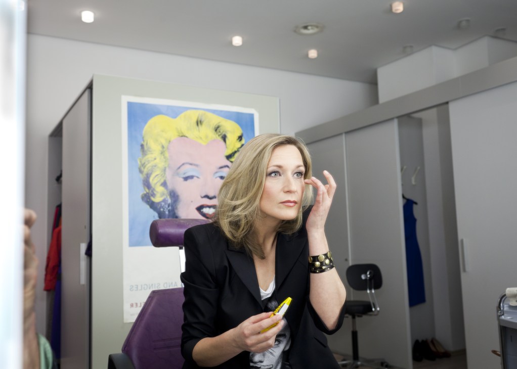 Presenter Meike Krüger in makeup, wearing a black blazer and gold bracelet, applying makeup while standing in front of a pop art portrait.