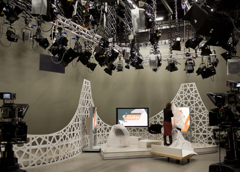 Deutsche Welle TV studio with abstract white geometric set design, professional lighting rigs overhead, and Arabic-language broadcast graphics on screens.