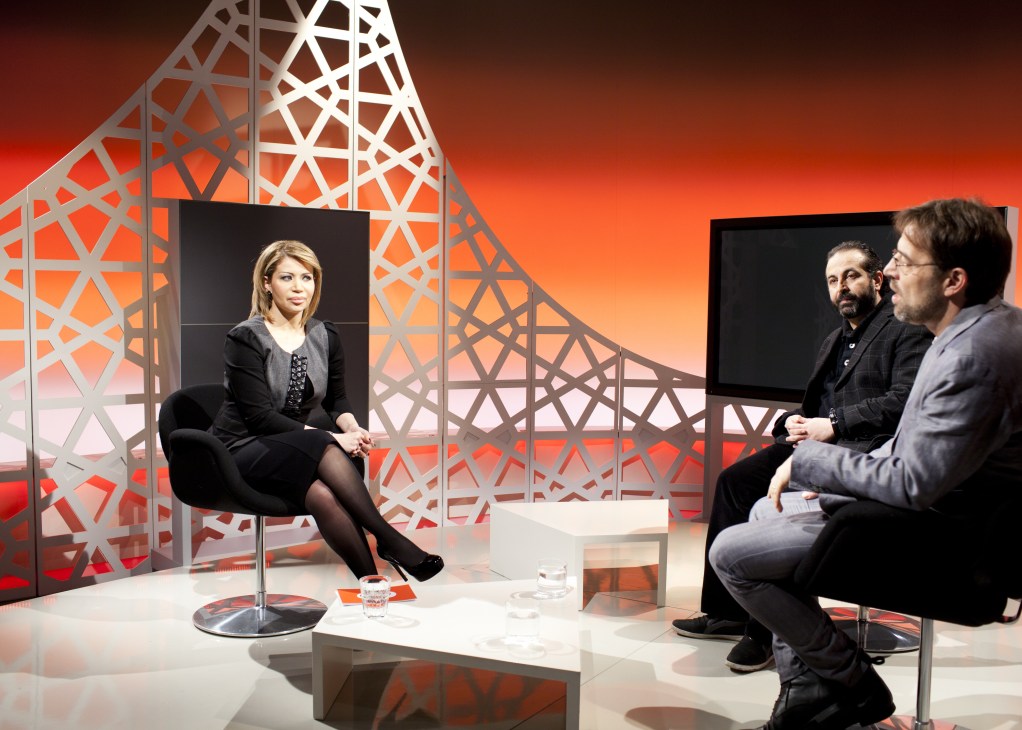 Presenter Dima Tarhini during a recording of On The Pulse talk show in Deutsche Welle's Berlin TV studio with red and orange abstract graphics.