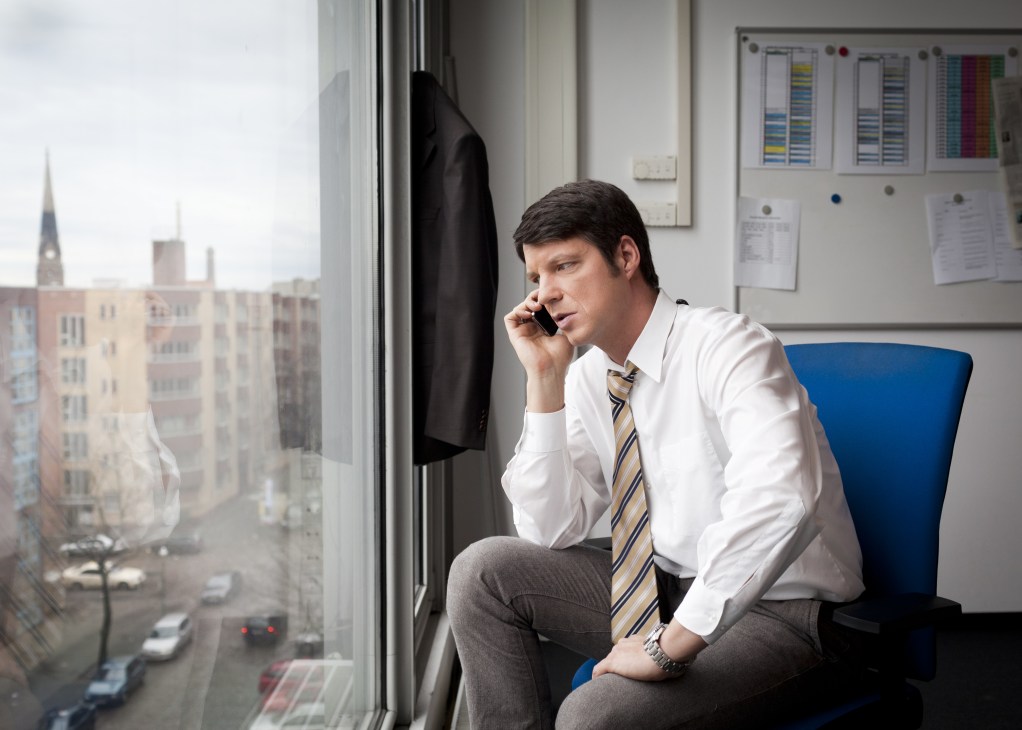News anchor Dirk Jacobs on a phone call in an office with a Berlin cityscape visible through the window.
