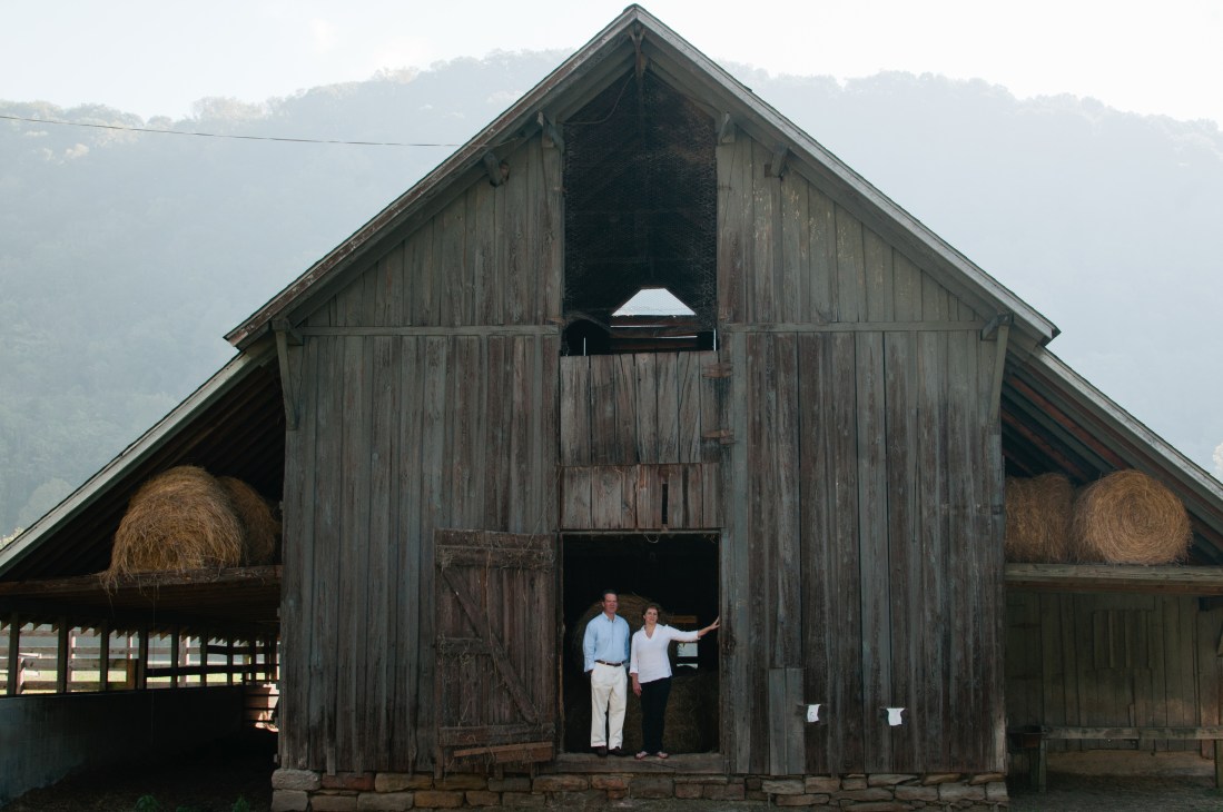 Two people stand in the doorway of a weathered wooden barn with hay bales visible inside and misty mountains behind.