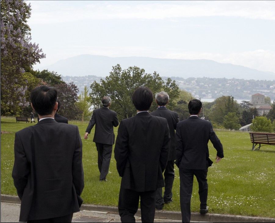 UN Asian delegation members in dark suits walking across a green park with Geneva's lake and mountains visible in the background.